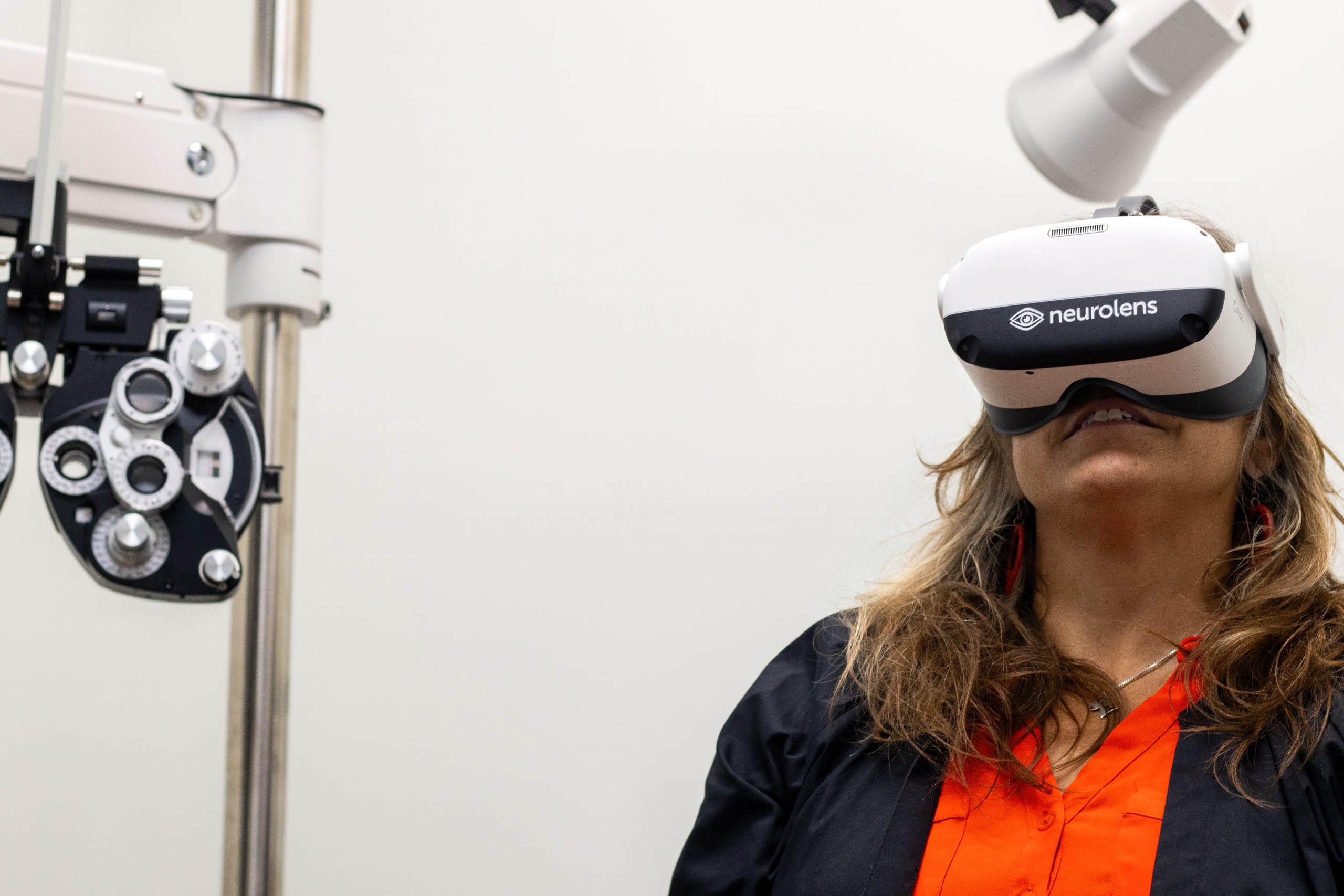 Image of smiling woman with long hair in the process of a Neurolens treatment. She is wearing the headgear and looking forward. The Neurolens logo is prominently displayed.