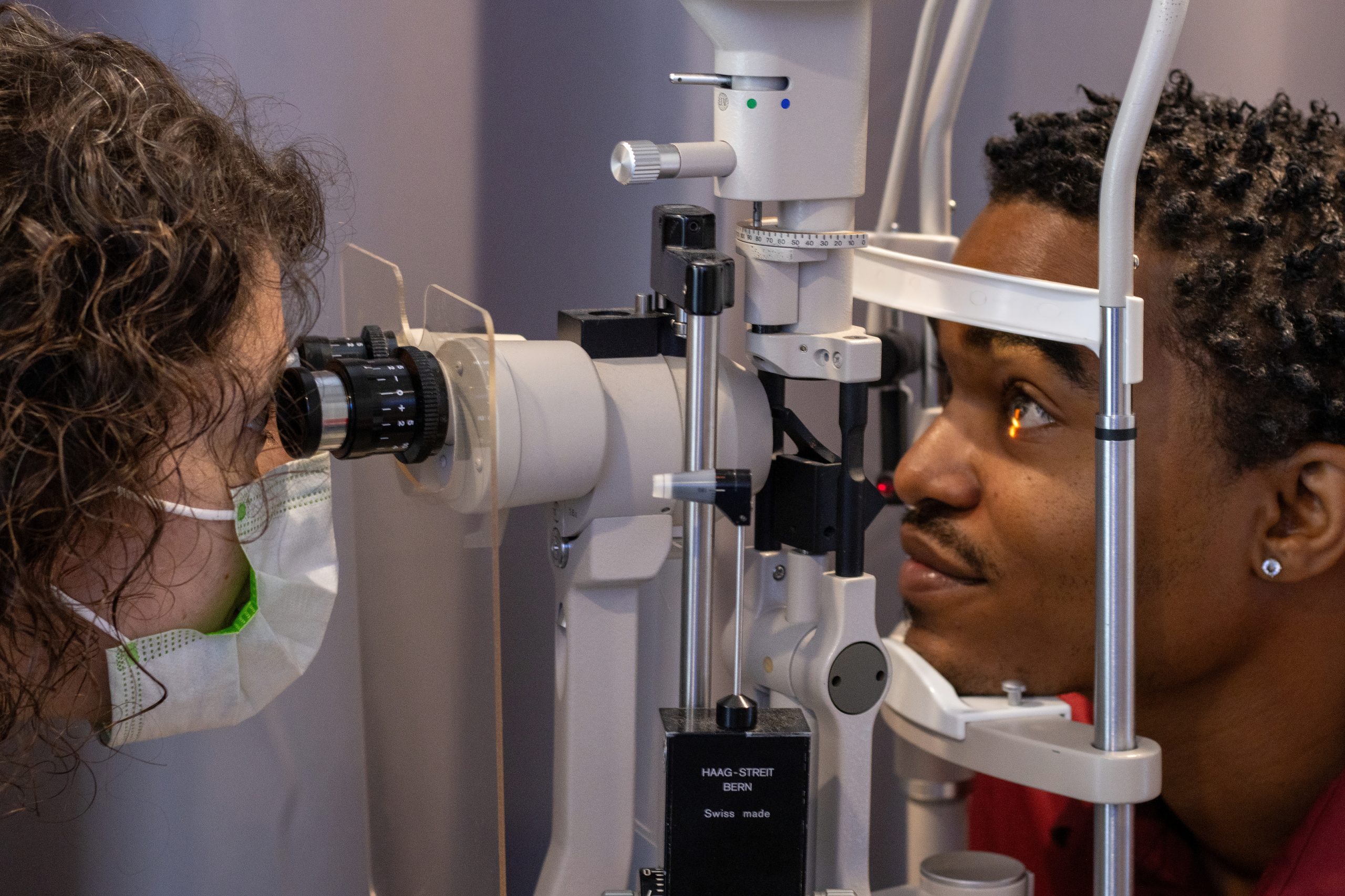 Image of an during an eye exam. His face is resting in eye care diagnostic equipment while an eye doctor peers through the eyepiece to examine the patient’s eye.