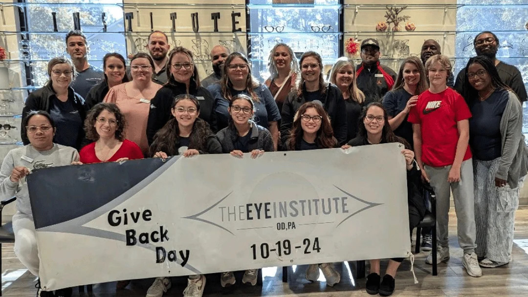 Group image from TEI Gives Back event. Smiling group members hold large banner displaying the event name and date.